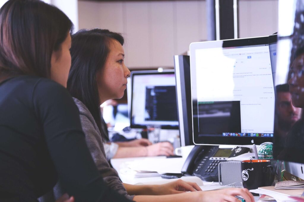 two women work at a mac computer in an open plan office