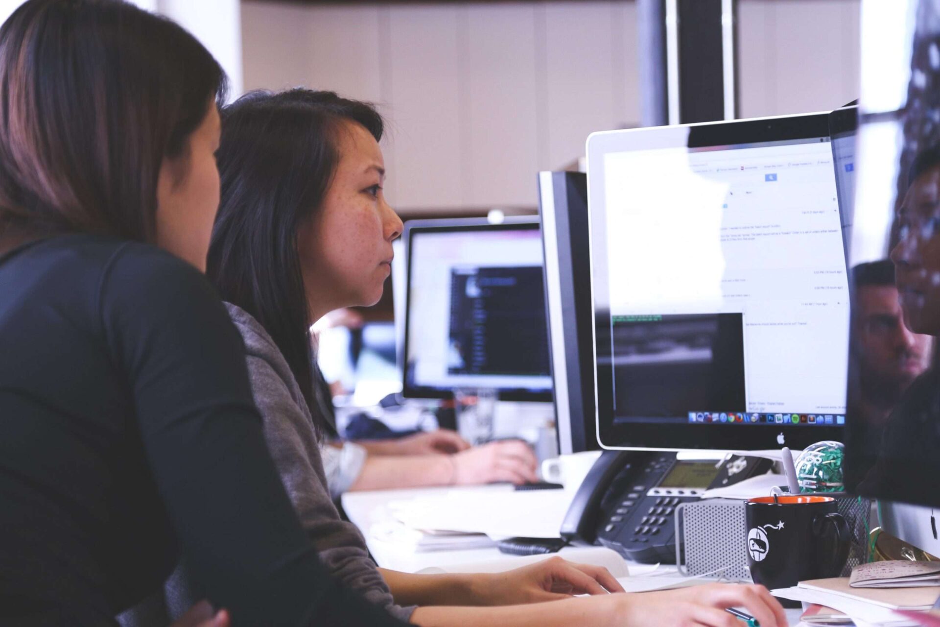 two women work at a mac computer in an open plan office