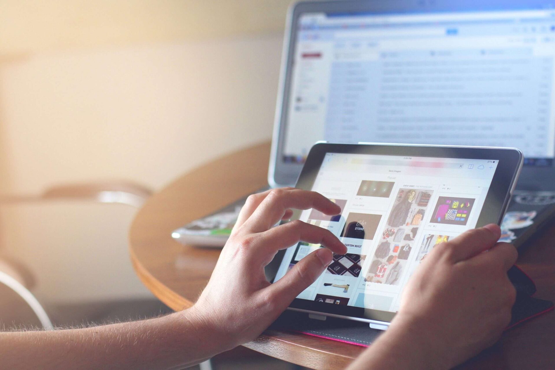 Close up of a hand browsing a site on their tablet with laptop open just behind