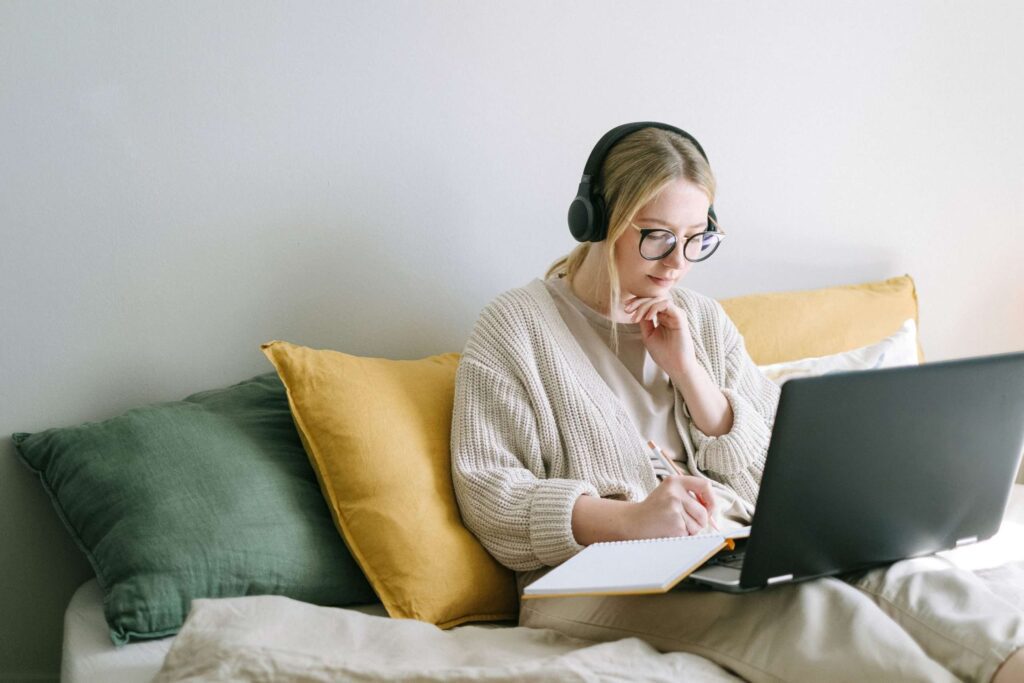 Woman using a laptop and wearing headphones takes notes in a notebook