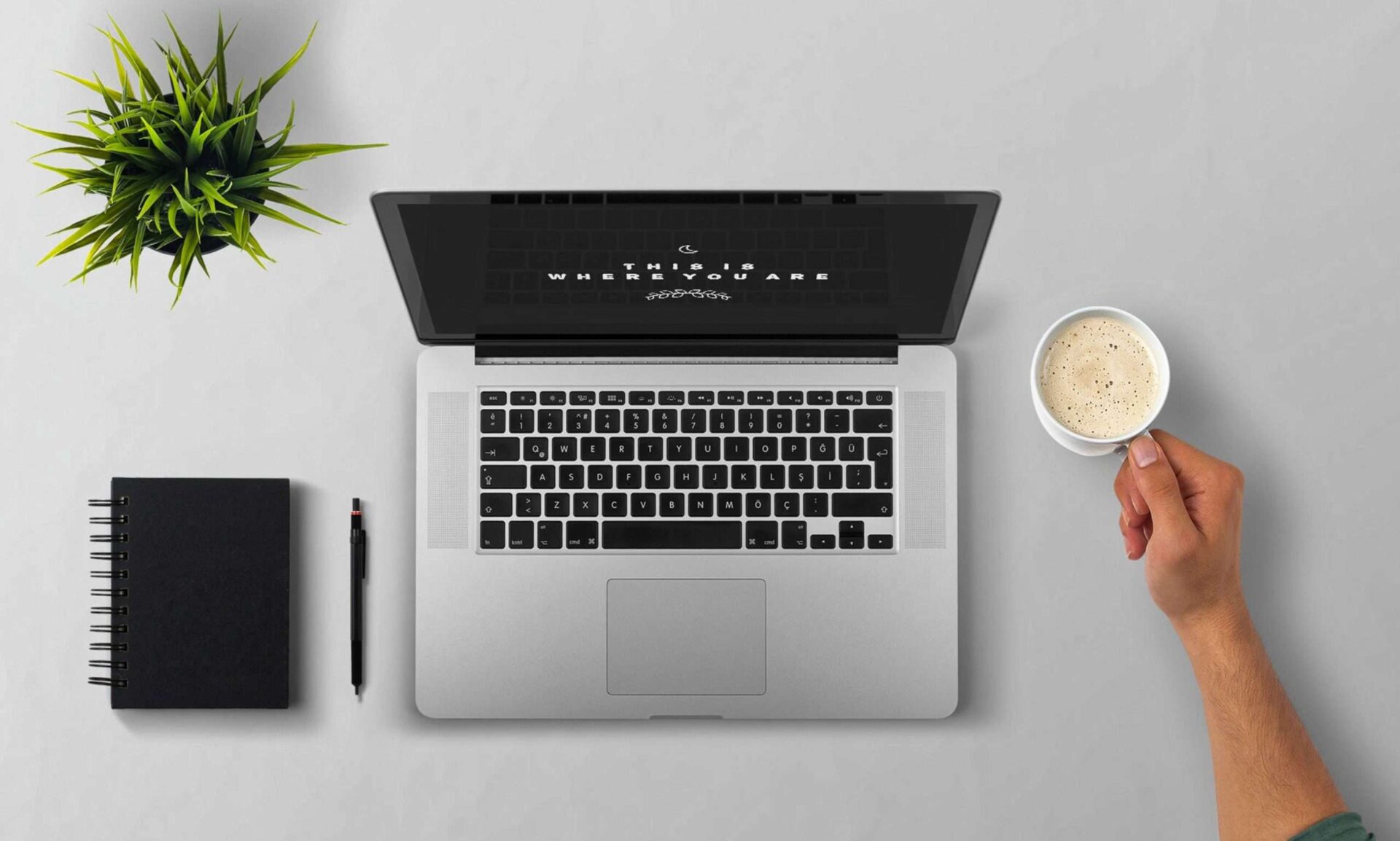 Top down view of a laptop and pad and pen with an arm holding a cup of coffee