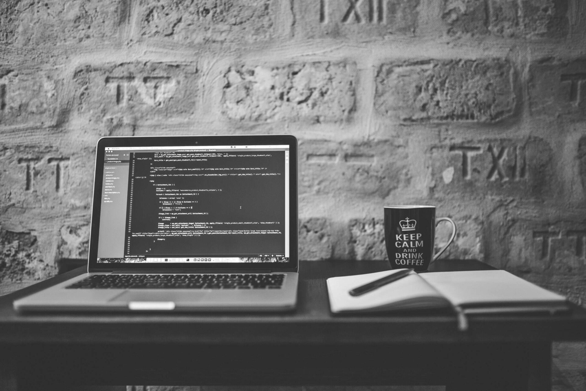 Greyscale photo of a laptop open next to a notebook and pen with a coffee mug that says Keep Calm and Drink Coffee