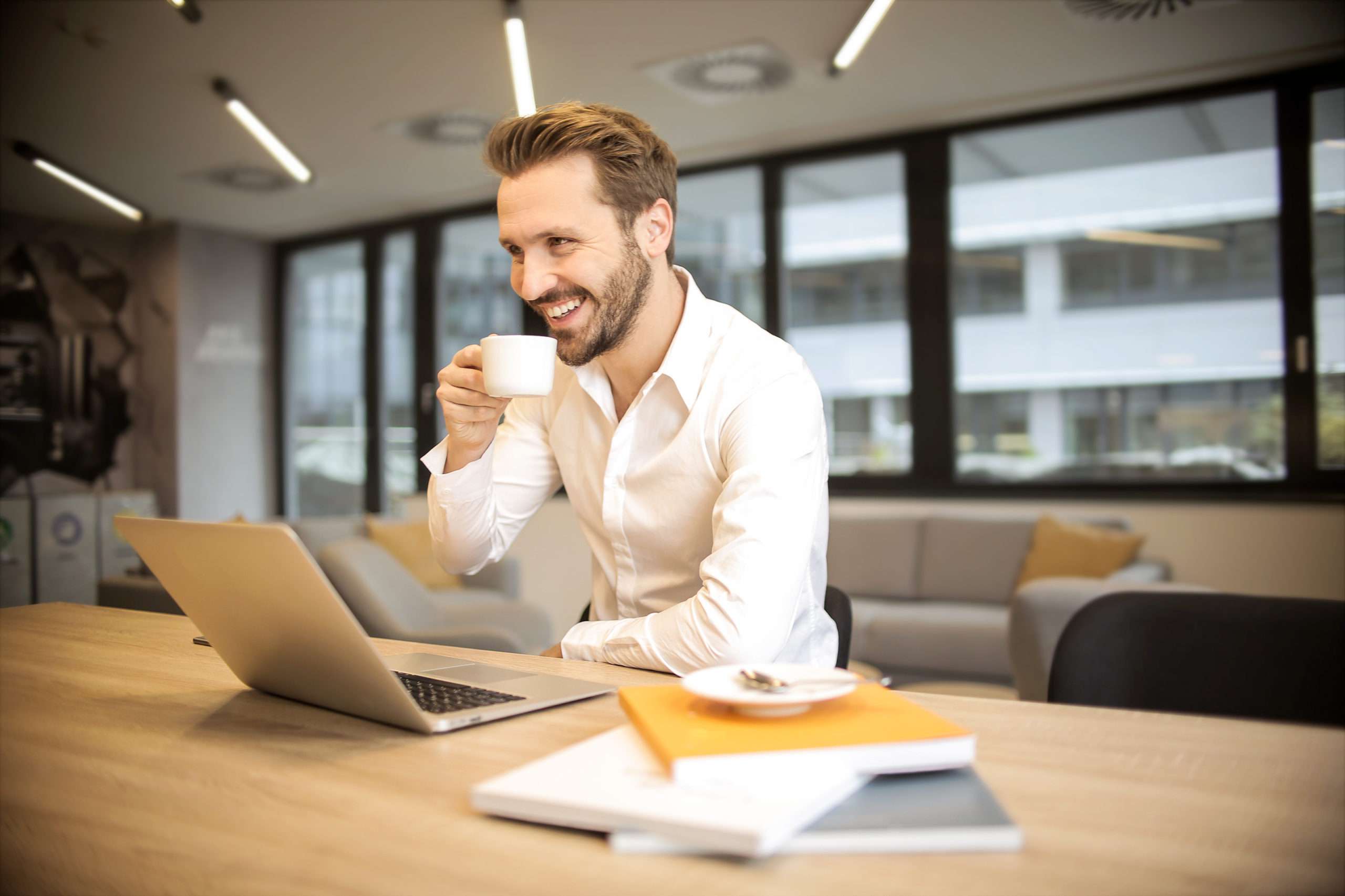 Smiling man drinking coffee working on a laptop in a modern office