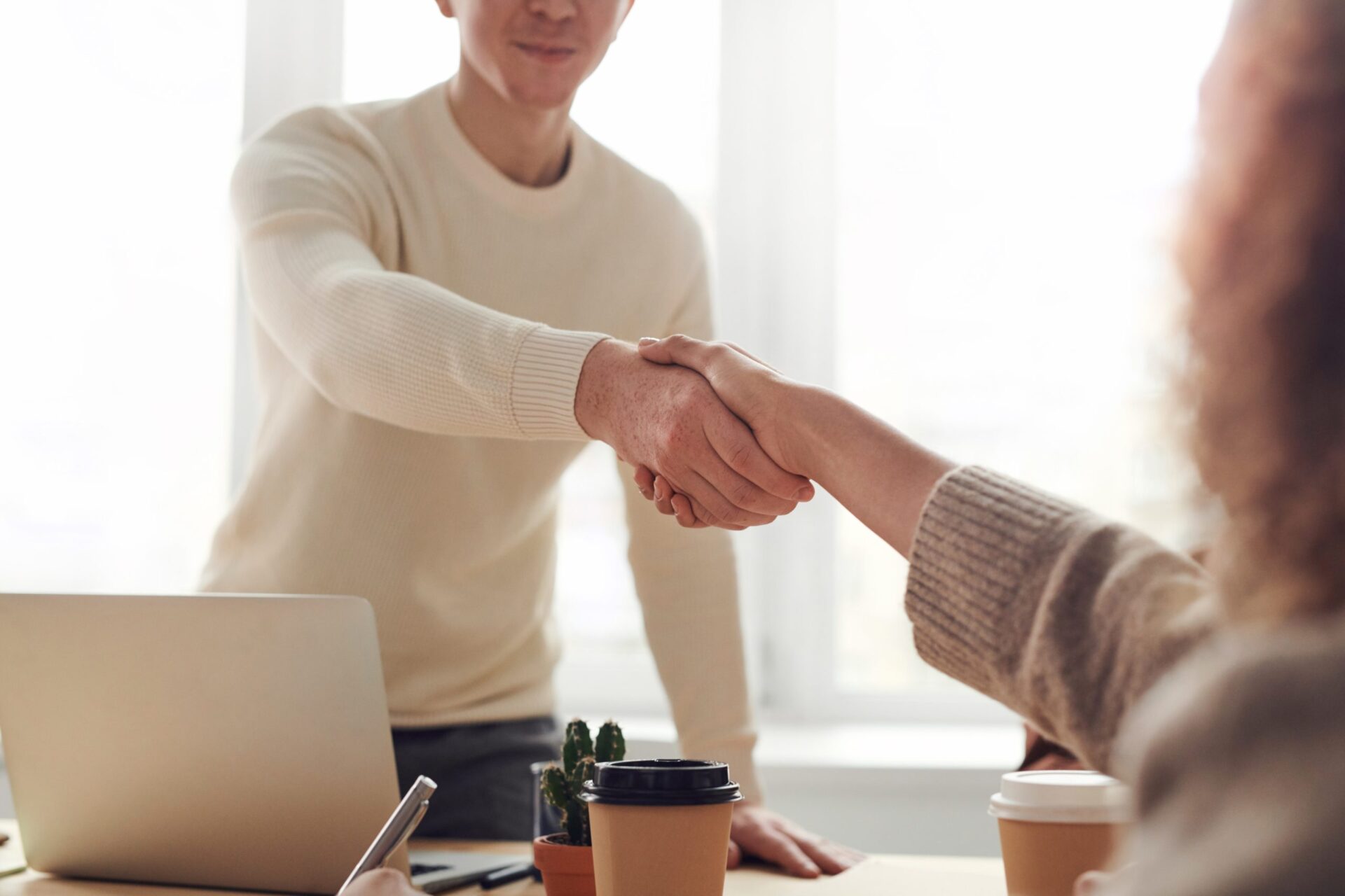 Two business people shaking hands over a desk with a laptop on