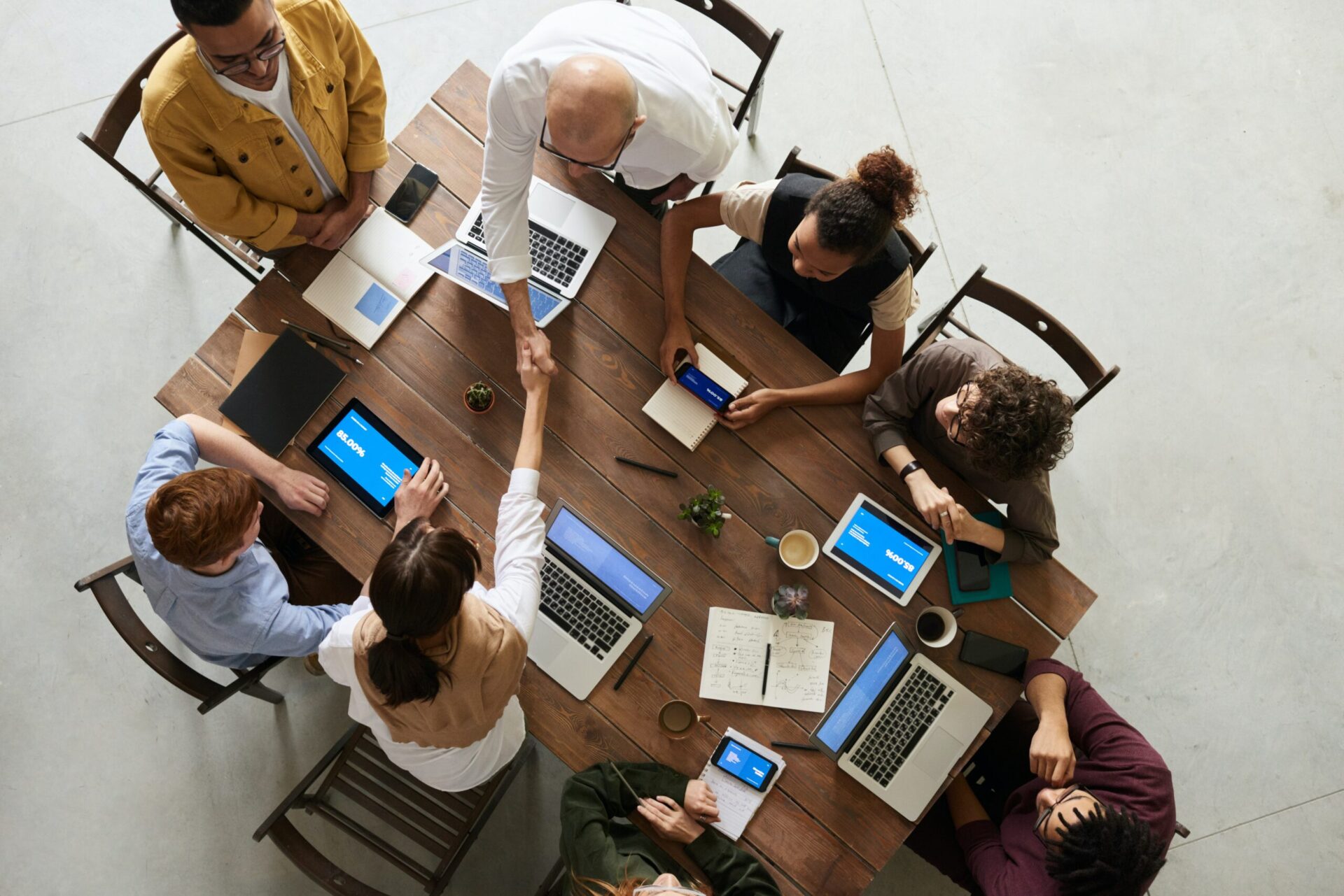 Topdown view of a several businesspeople meeting around a table that is full of tablets and laptops as two people on either side of the table shake hands