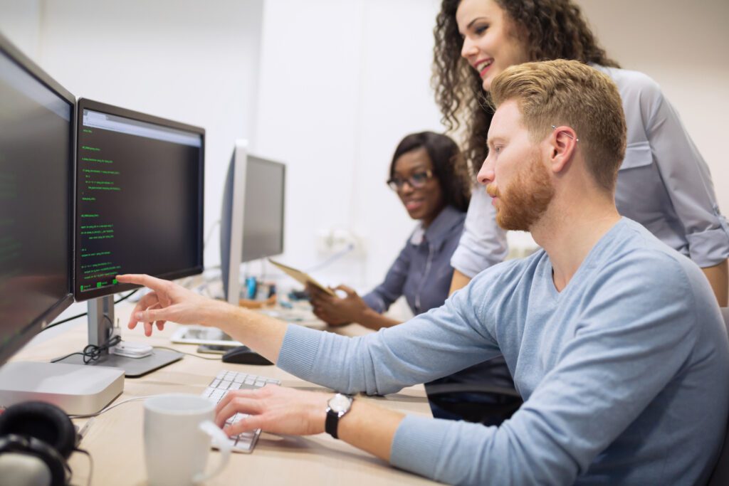 Programmer working at a computer with multiple screens as two coworkers look as he points to a line of code on screen
