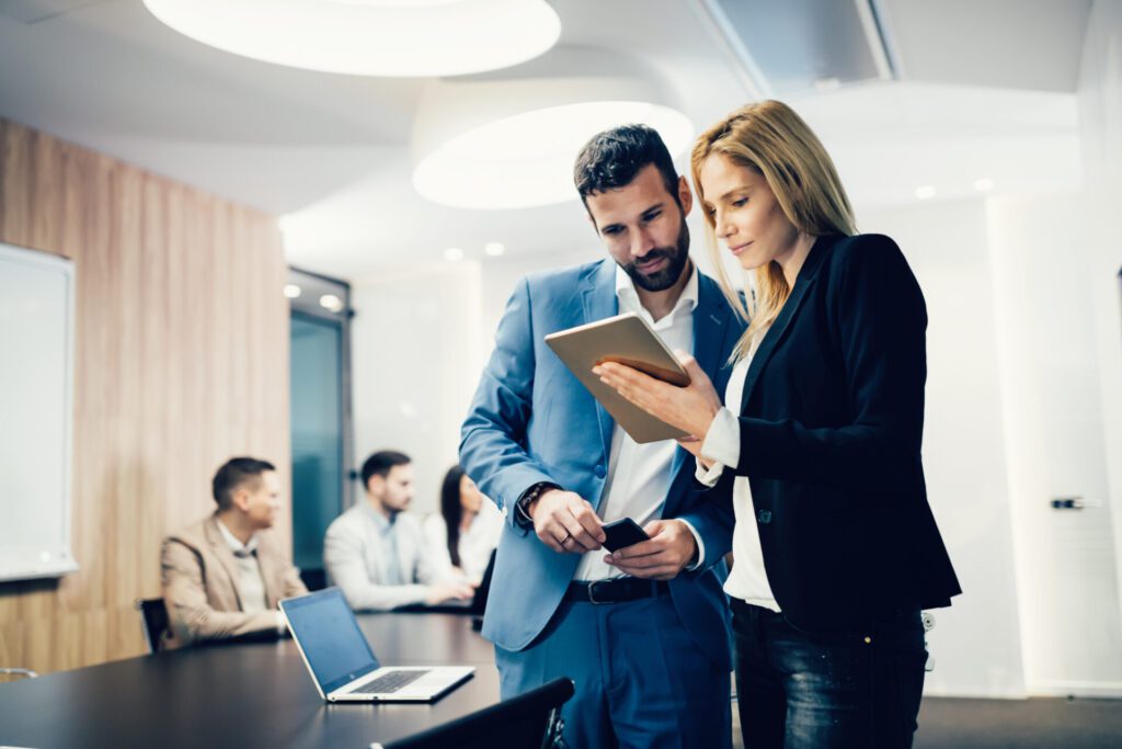 Two business colleagues looking at something on a tablet as other people meet around a table in the background