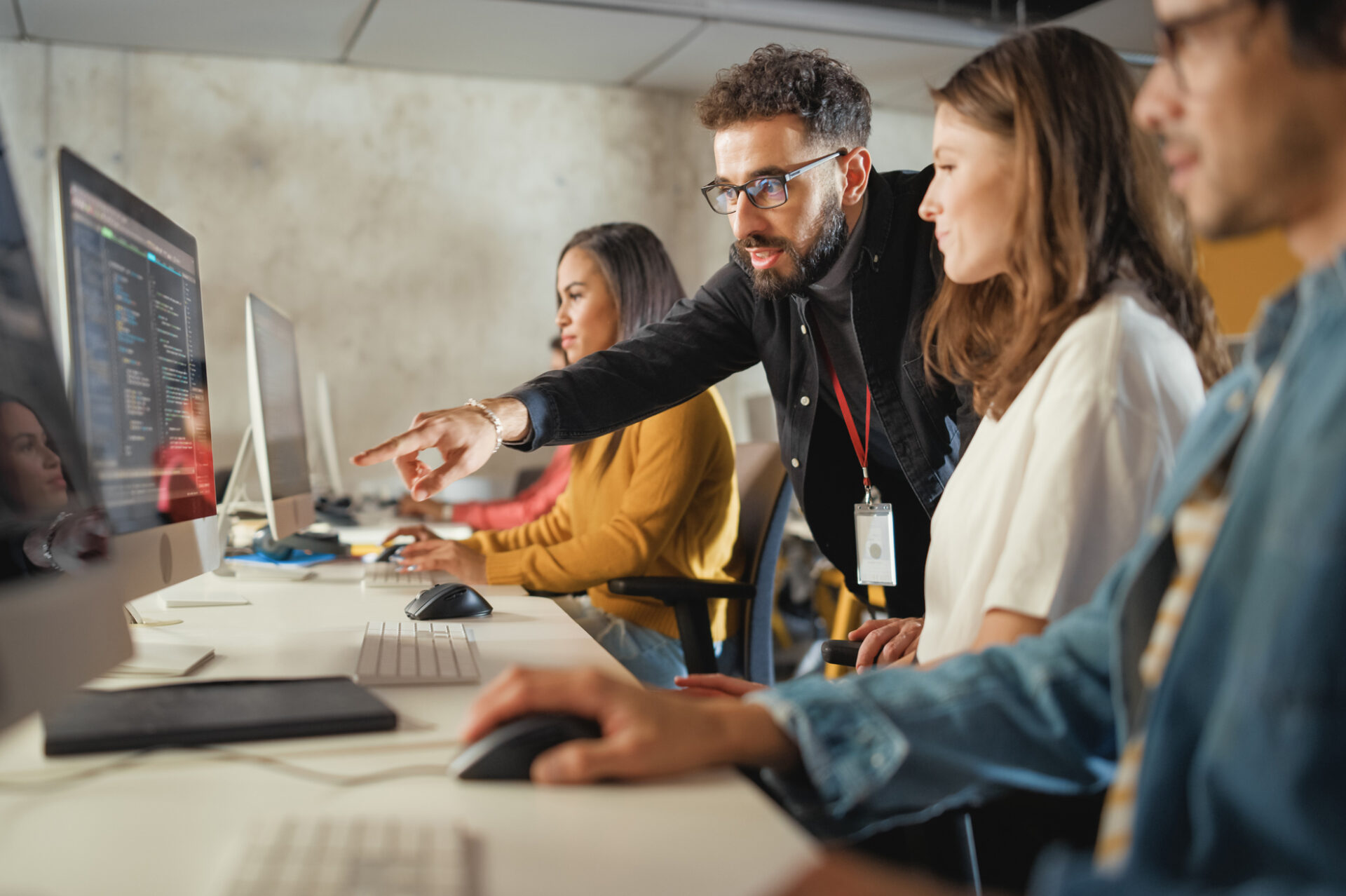 Group of people working at mac computers as a teacher points to something on one of the screens