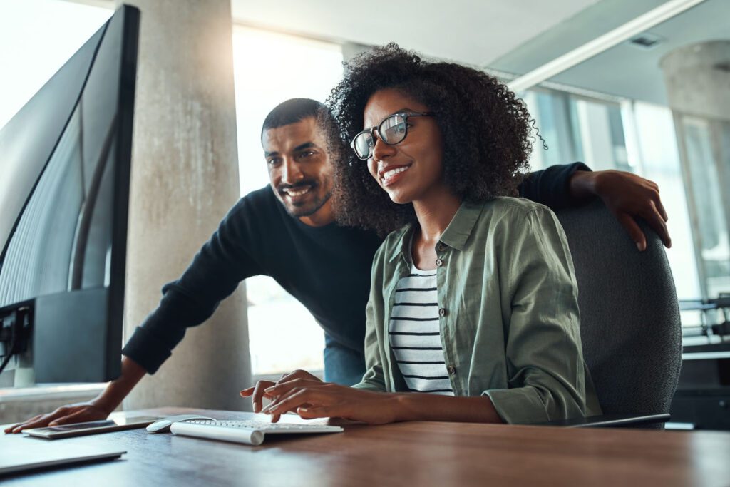 Two business professionals smiling and looking at a desktop computer as they work in an open plan office