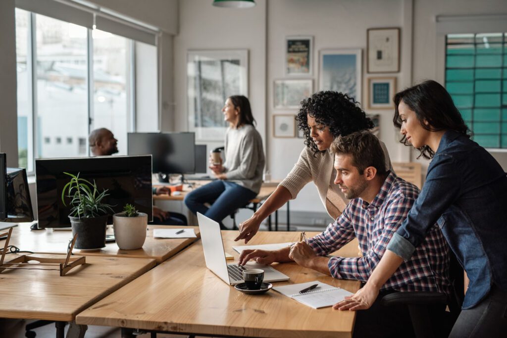 An open plan office with several people looking at a computer offering advice to the user