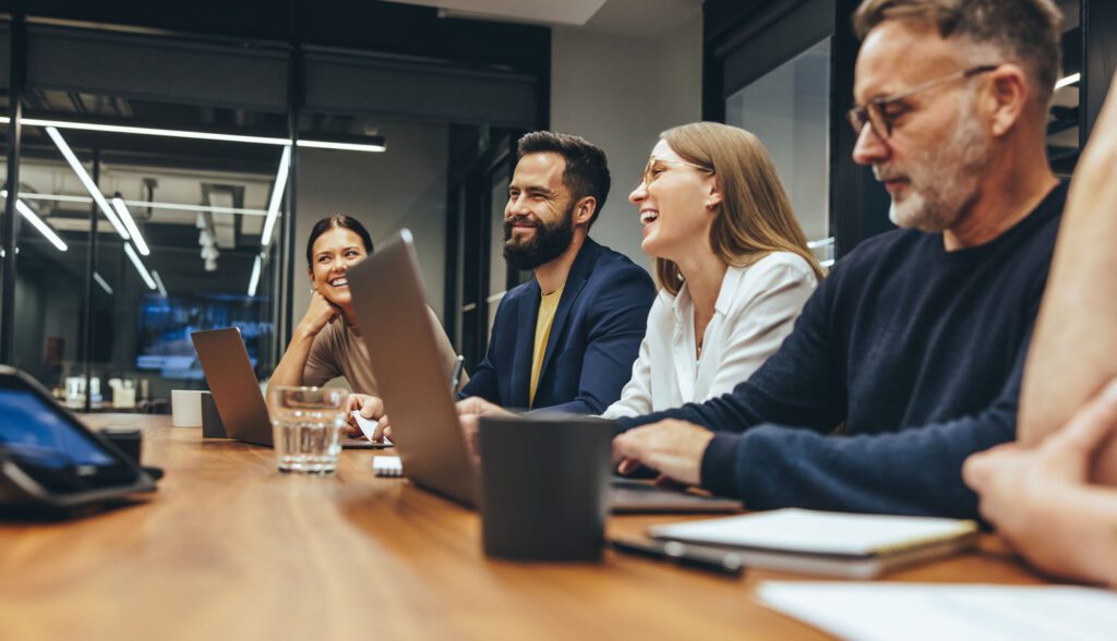 Bunch of business people around a table smiling and talking as they use laptops