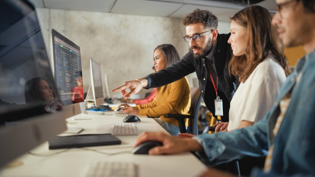 Group of people working at mac computers as a teacher points to something on one of the screens