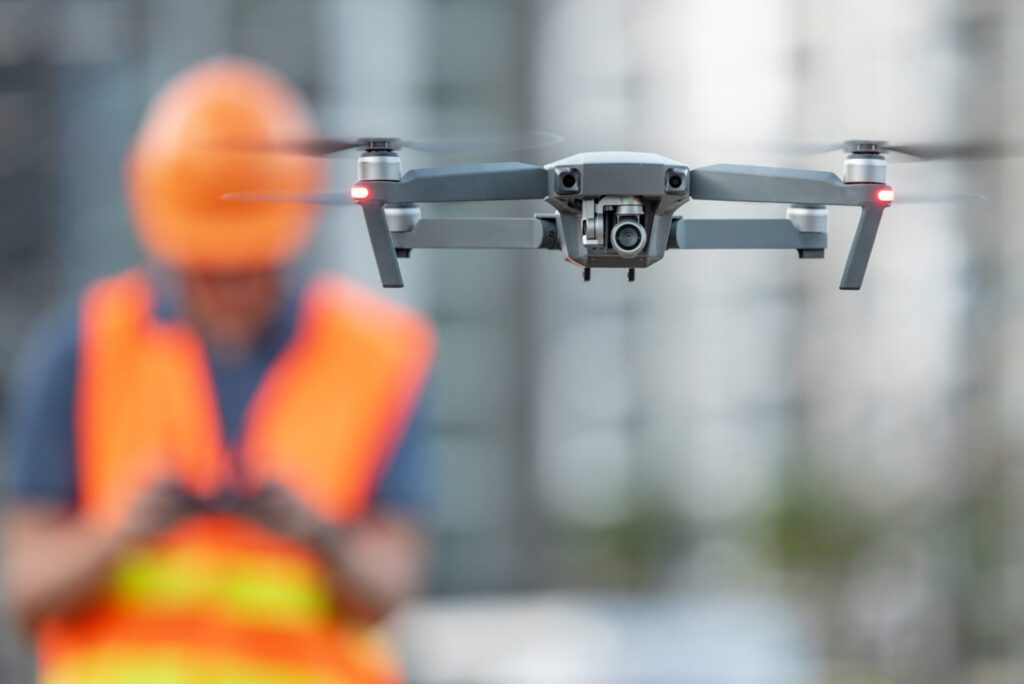 Close up photo of a drone flying with an out of focus man in hi vis jacket and hard hat behind