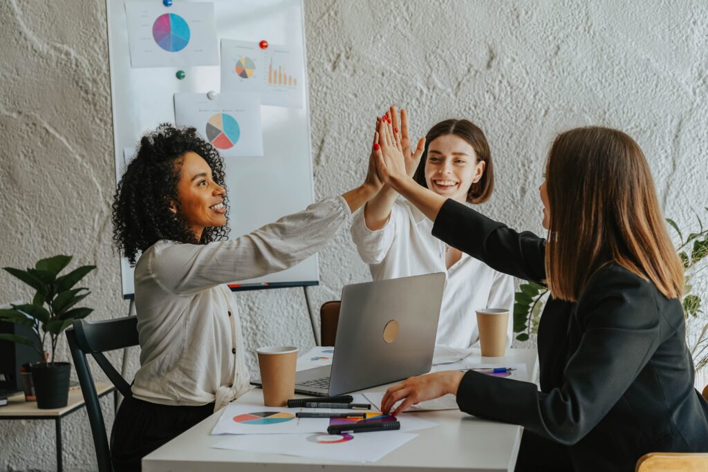 Three businesswomen hi-fiving as they sit around a laptop in an office environment
