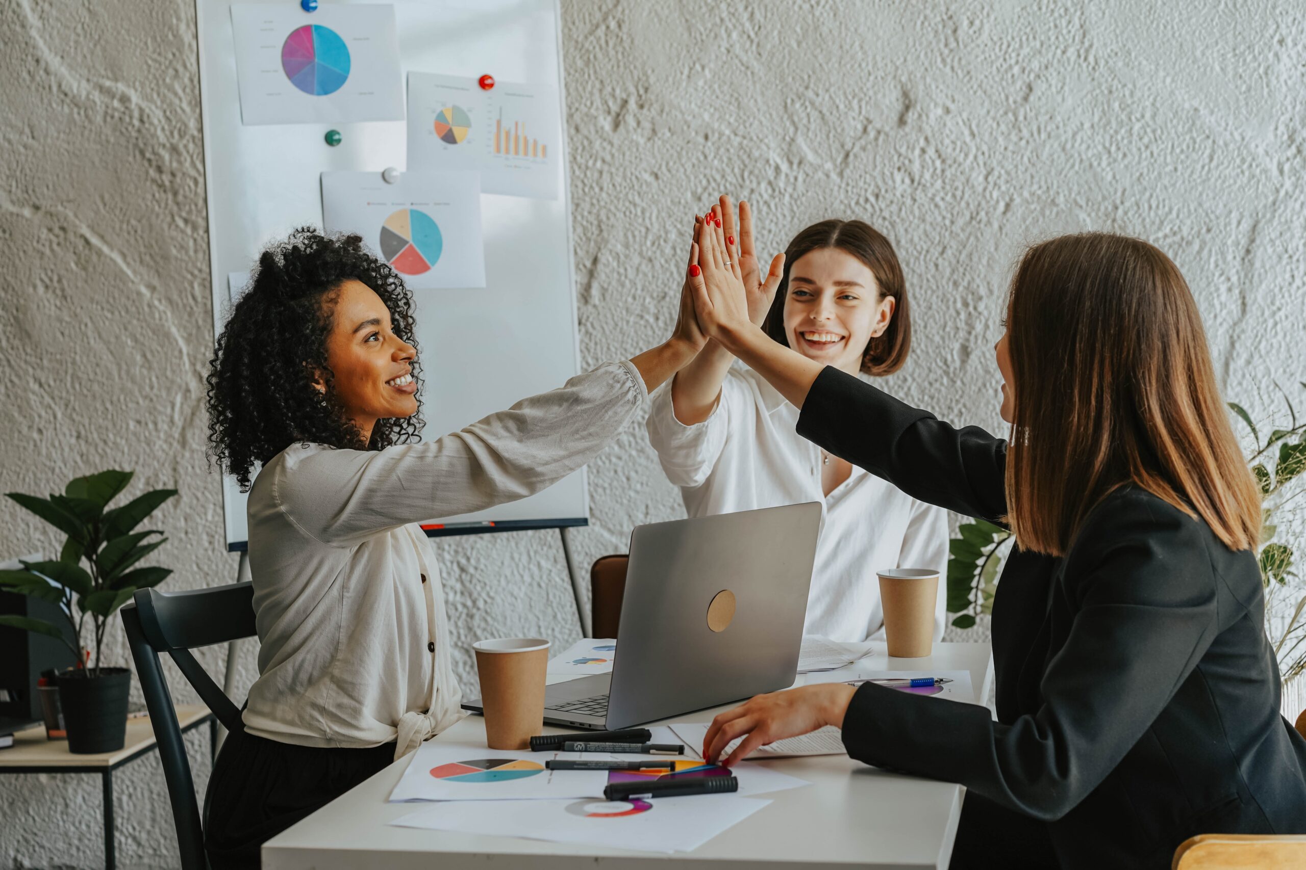Three businesswomen hi-fiving as they sit around a laptop in an office environment