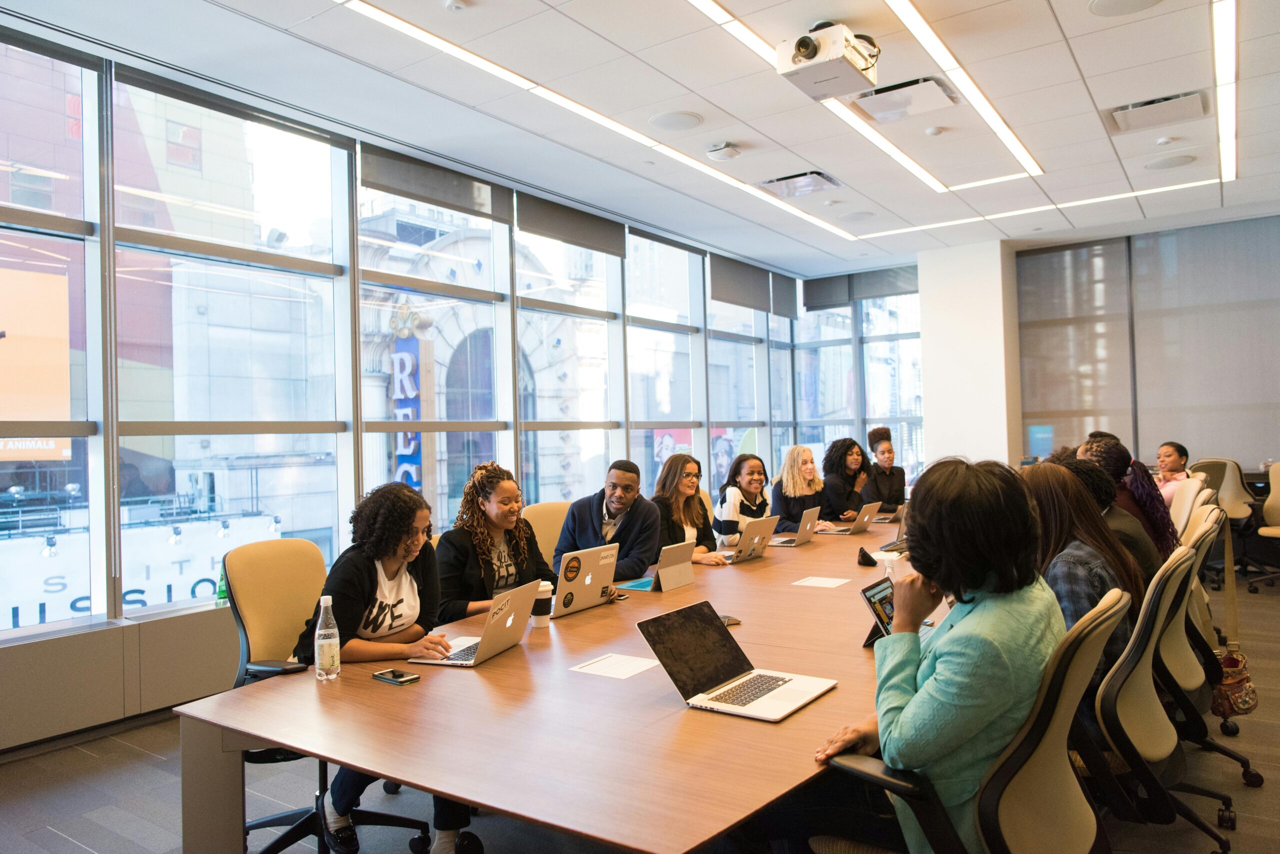 Wide photo of a large meeting table surrounded by business people talking and working at laptops