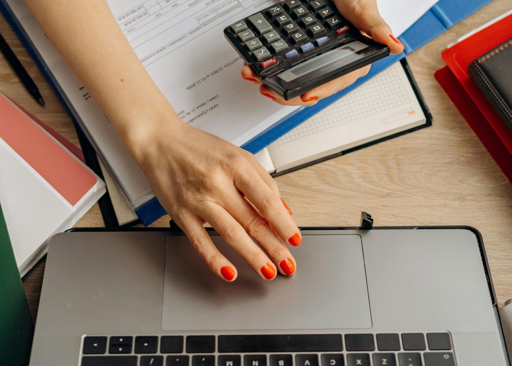 Closeup of a person using a calculator as they work at a laptop on a desk filled with papers and books