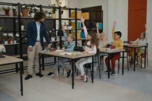 Photo inside a primary school with a teacher asking a question and all the students holding their hand up to answer