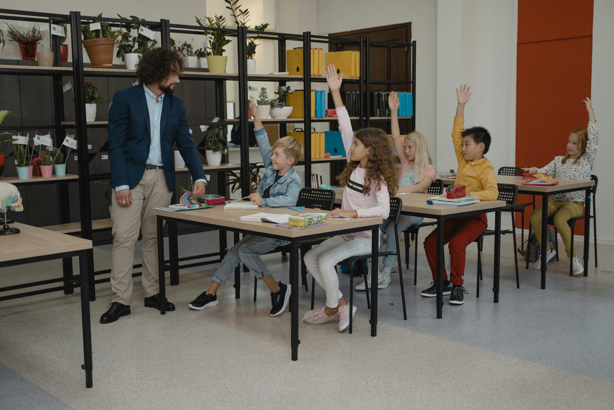 Photo inside a primary school with a teacher asking a question and all the students holding their hand up to answer