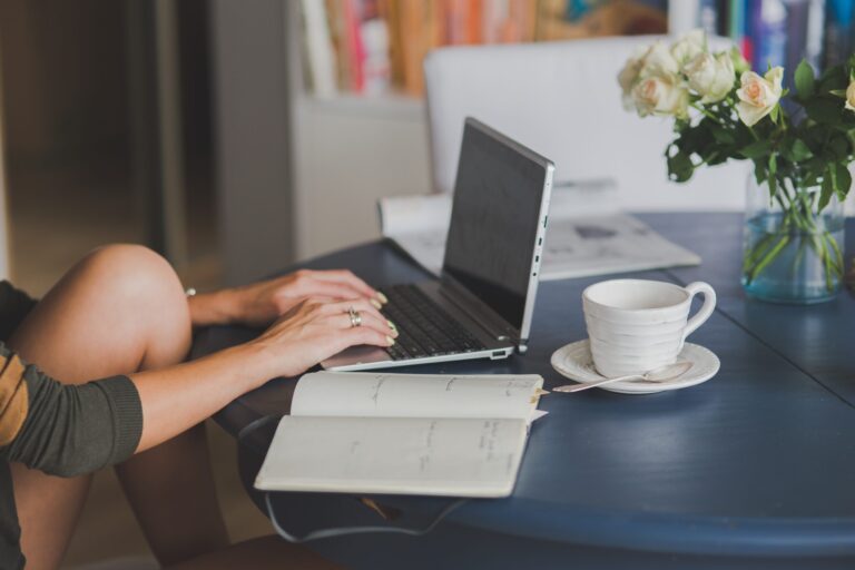 Person typing at a laptop on a table with an empty coffee mug and open notepad nearby