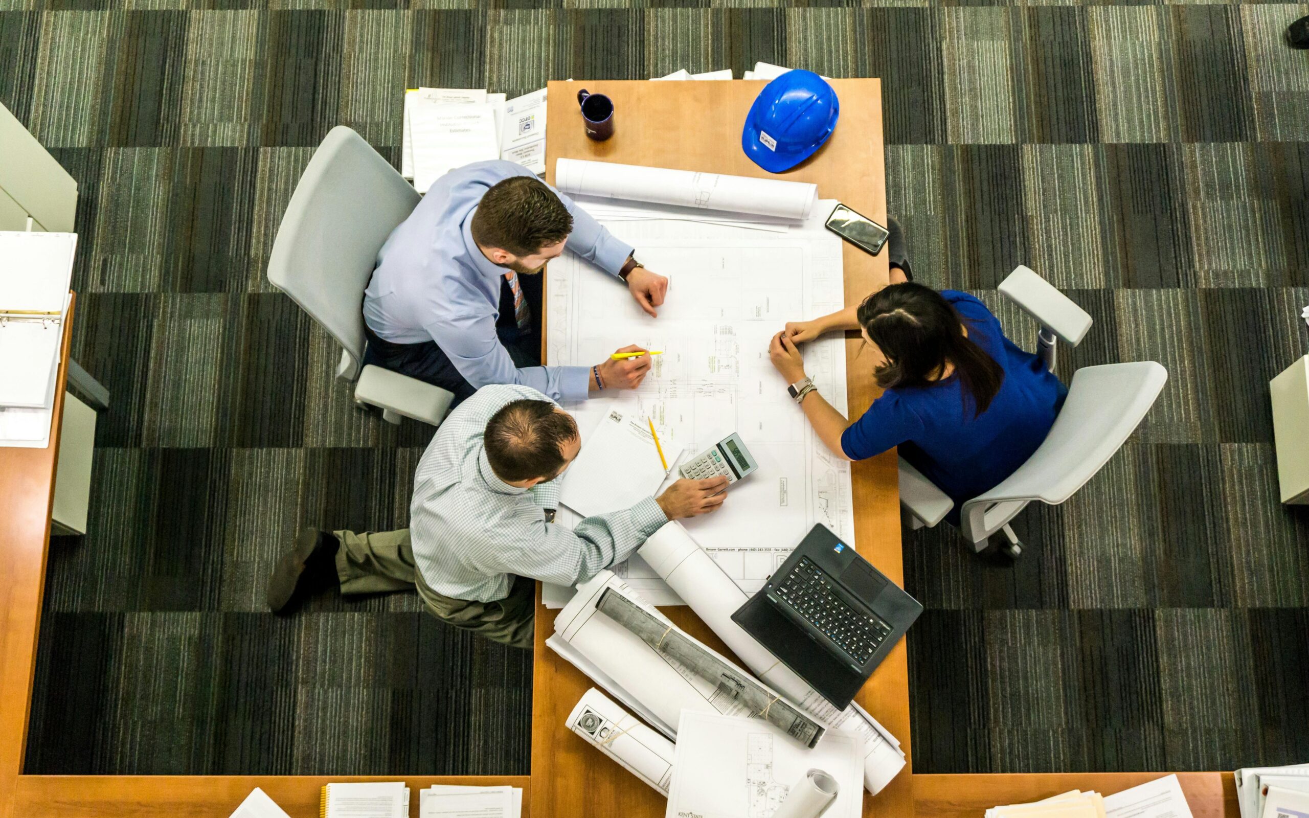 Top down view of several people working at a desk with a laptop and computer as well as construction schematics and plans