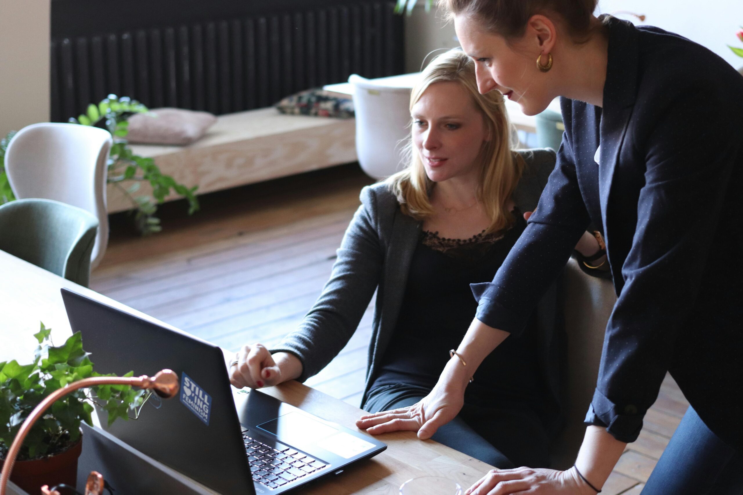 Two women talking about something on a laptop on a desk in a casual office setting