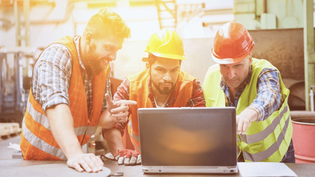 Three construction workers in hi-vis jackets and hard hats stand by a table on a construction site looking at a laptop.