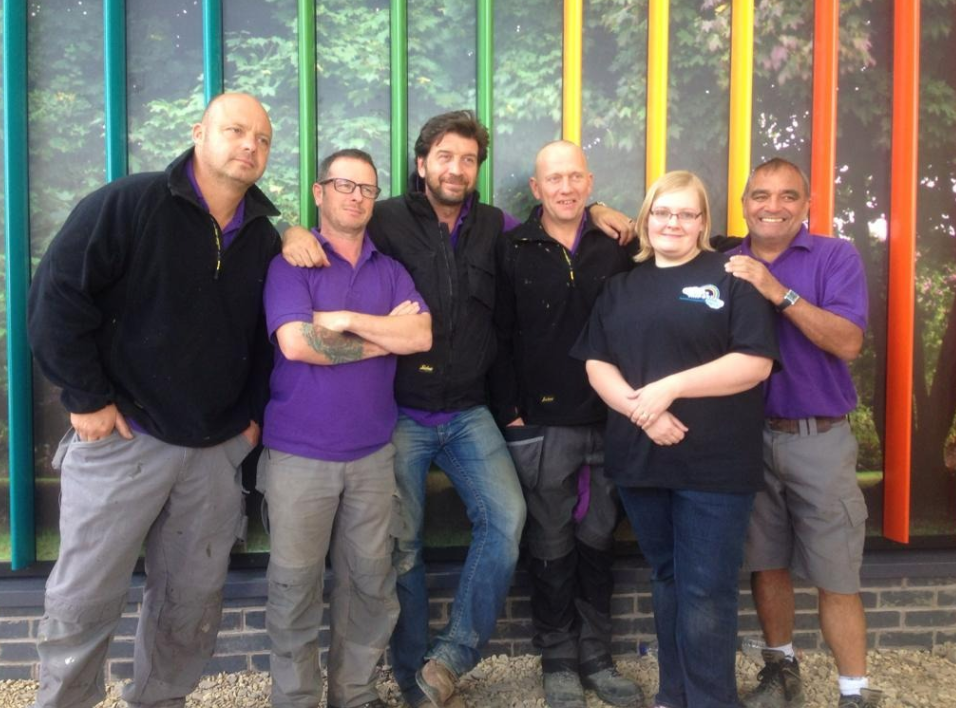 A group of five middle-aged men and a middle-aged woman dressed in Little Miracles’ charity colours stand in front of a wall. The wall is covered with a large image of a tree and is divided up by rainbow-coloured panels that transition from light blue on the left to orange on the right.