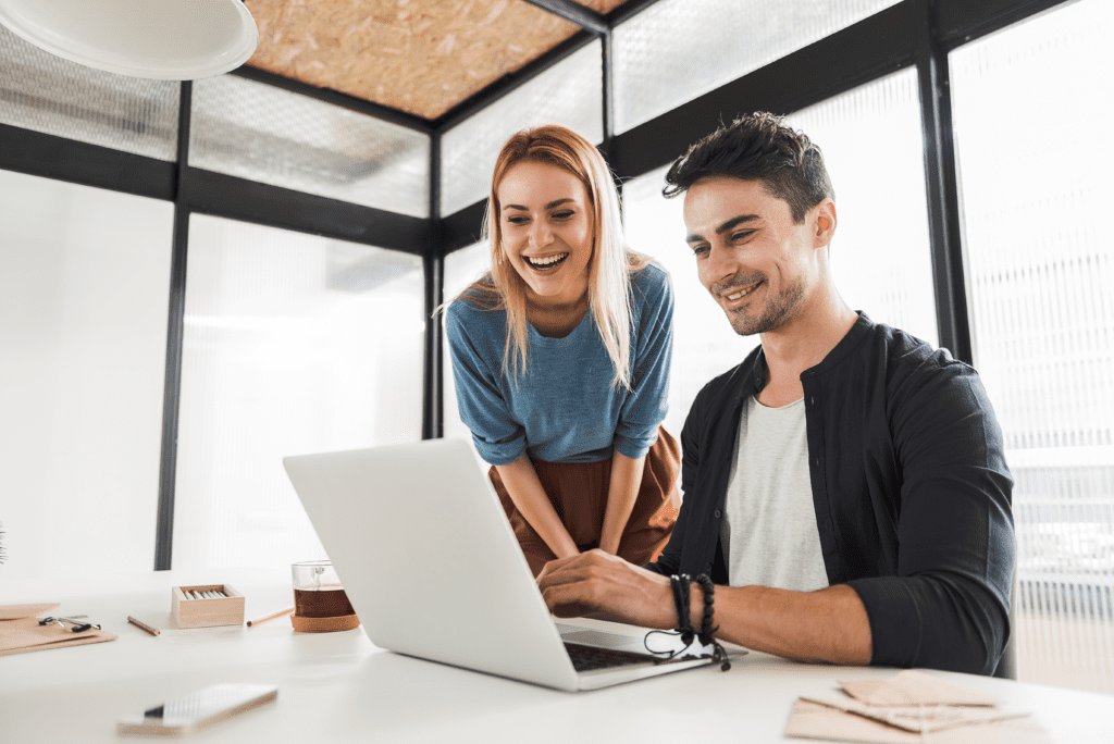 A young, casually dressed, smiling businesswoman and businessman stand in front of a laptop.