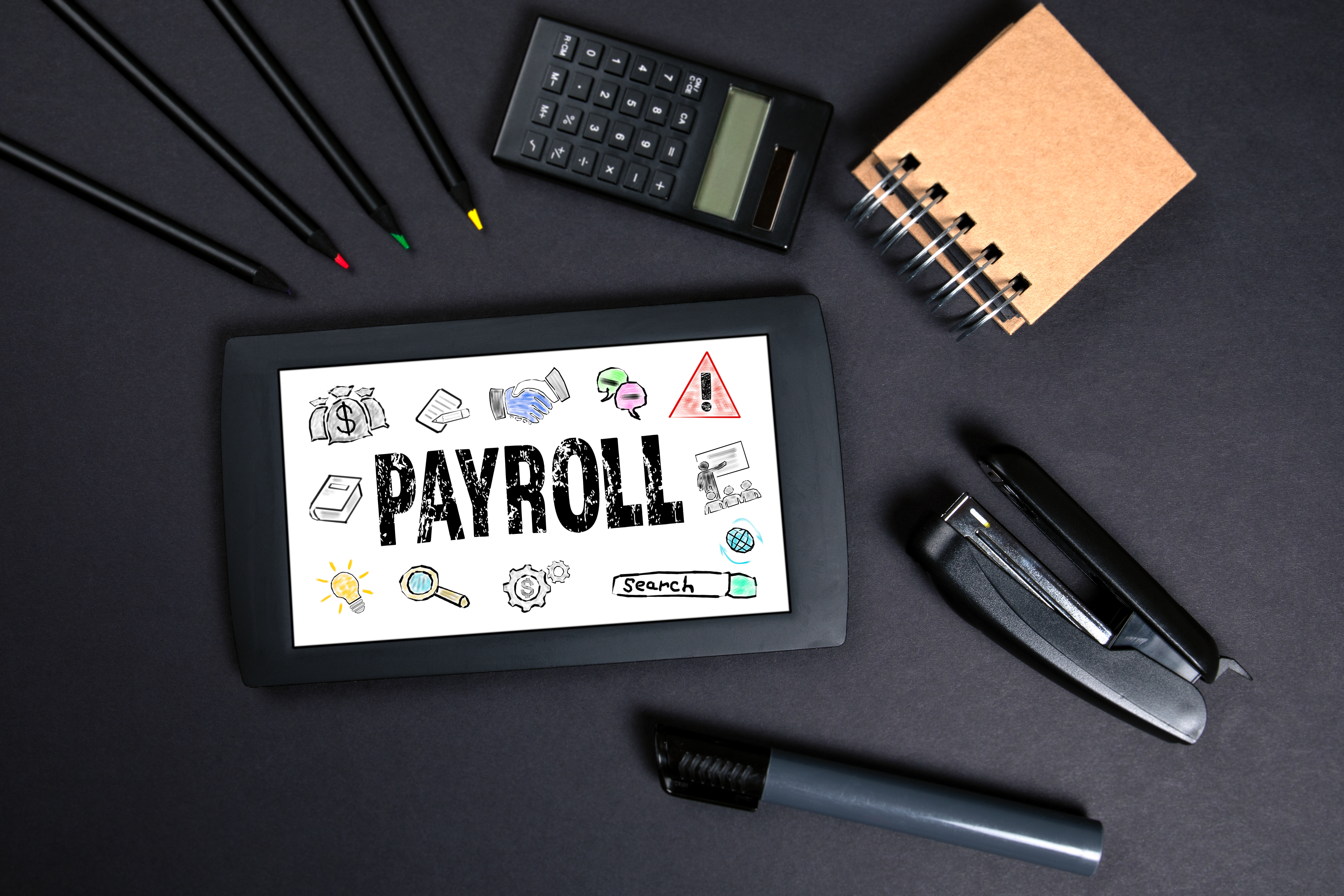An image showing pencils; a calculator; a notebook; a stapler; a whiteboard pen; and a screen on a black paper background. The screen says “PAYROLL” with doodles, such as a bag of money and a lightbulb, around it.