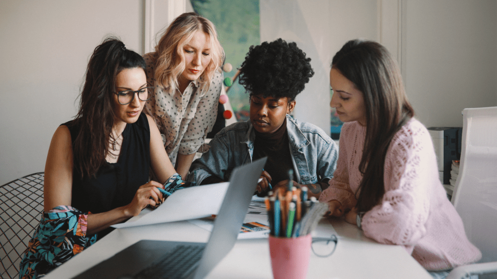 Four young women sit in an office space around a laptop.