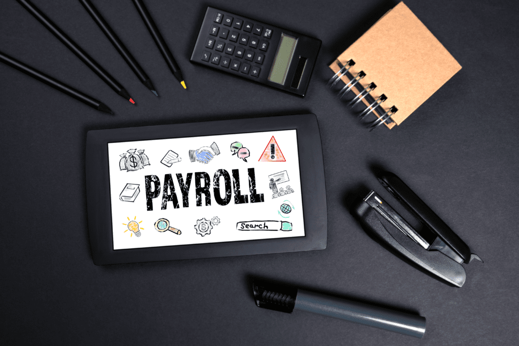 An image showing pencils; a calculator; a notebook; a stapler; a whiteboard pen; and a screen on a black paper background. The screen says “PAYROLL” with doodles, such as a bag of money and a lightbulb, around it.