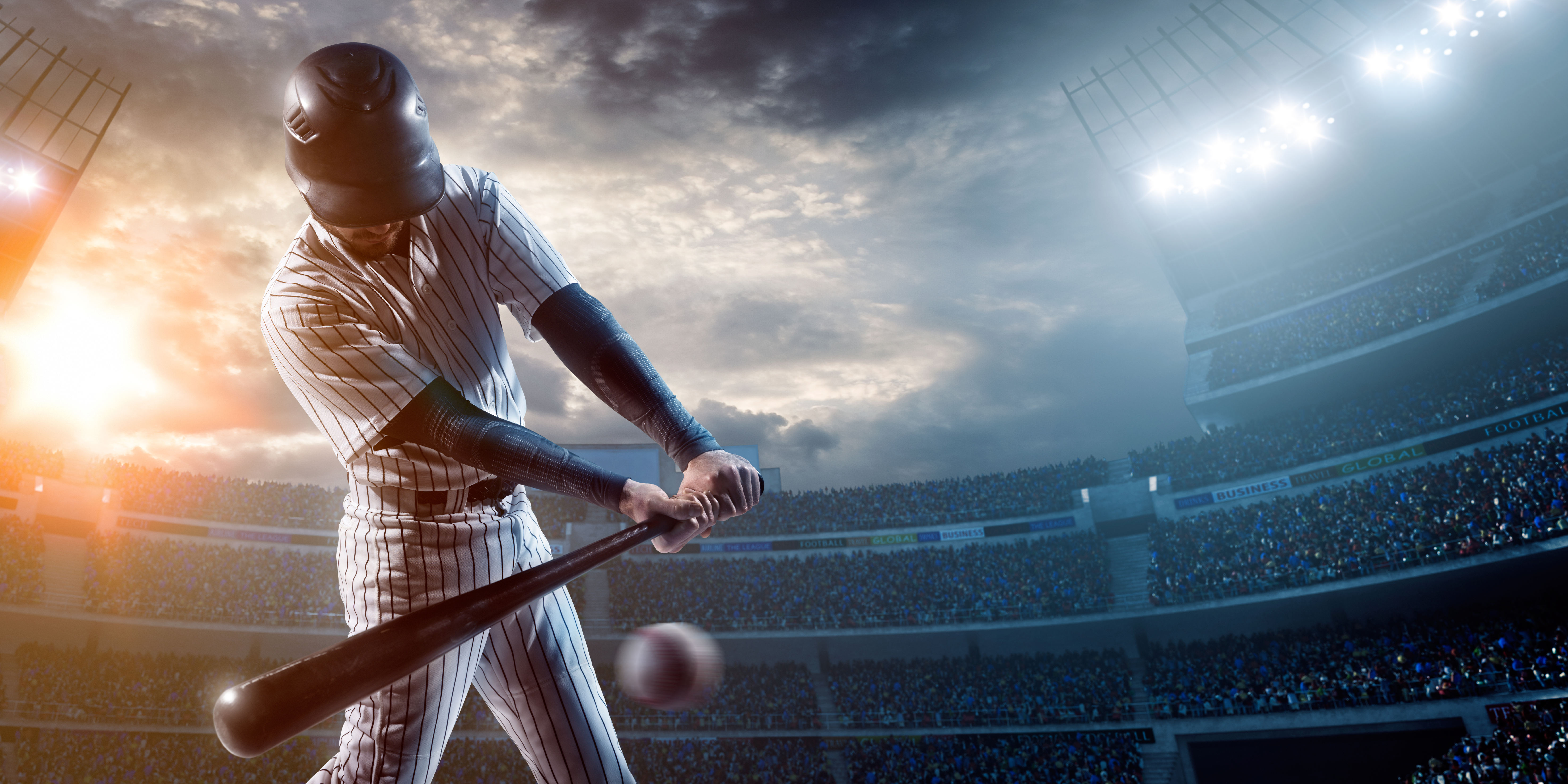 A baseball player swings his bat at a baseball in a busy stadium at sunset.