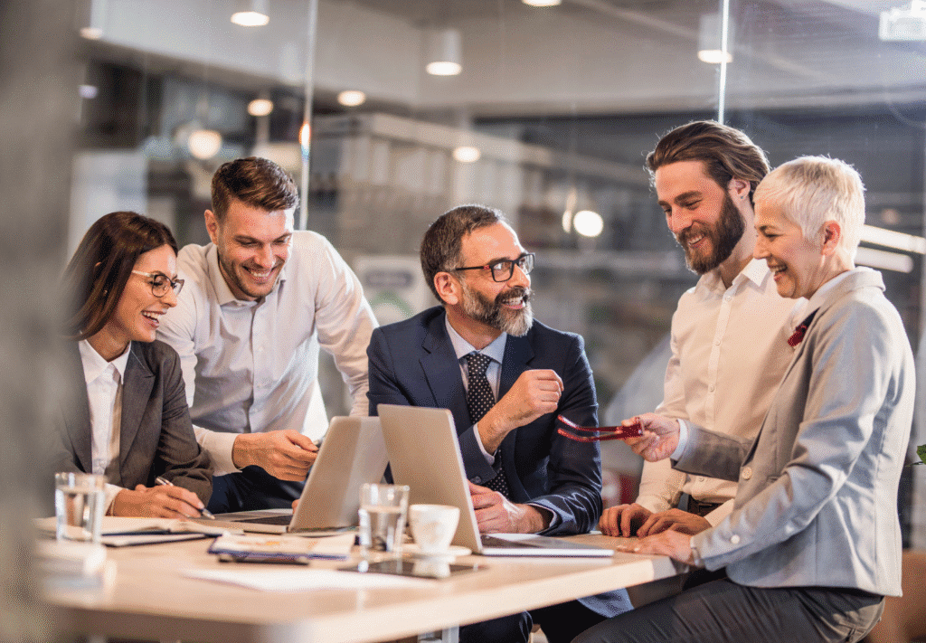 A group of smiling businesspeople stand around a desk with two laptop computers.