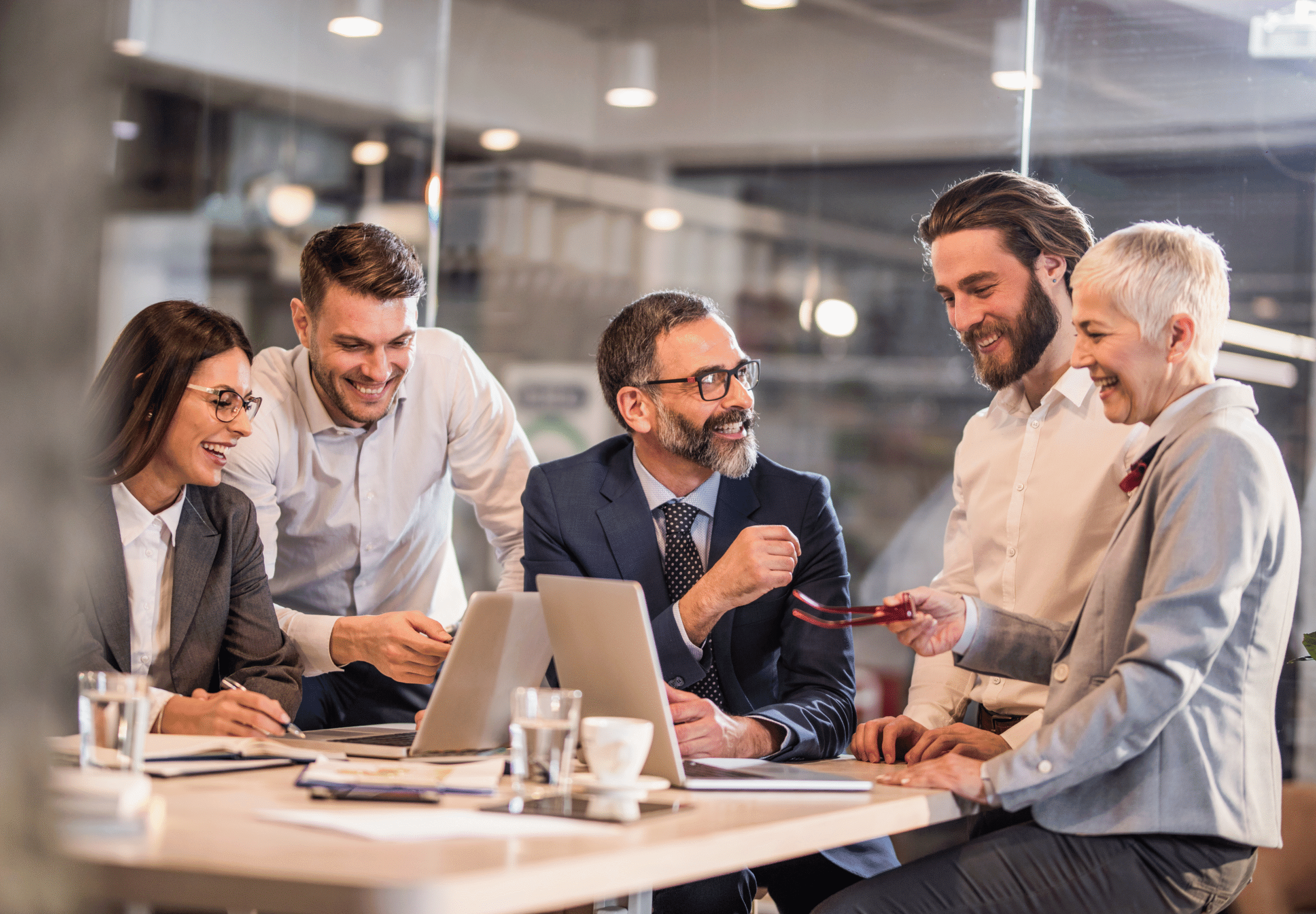 A group of smiling businesspeople stand around a desk with two laptop computers.