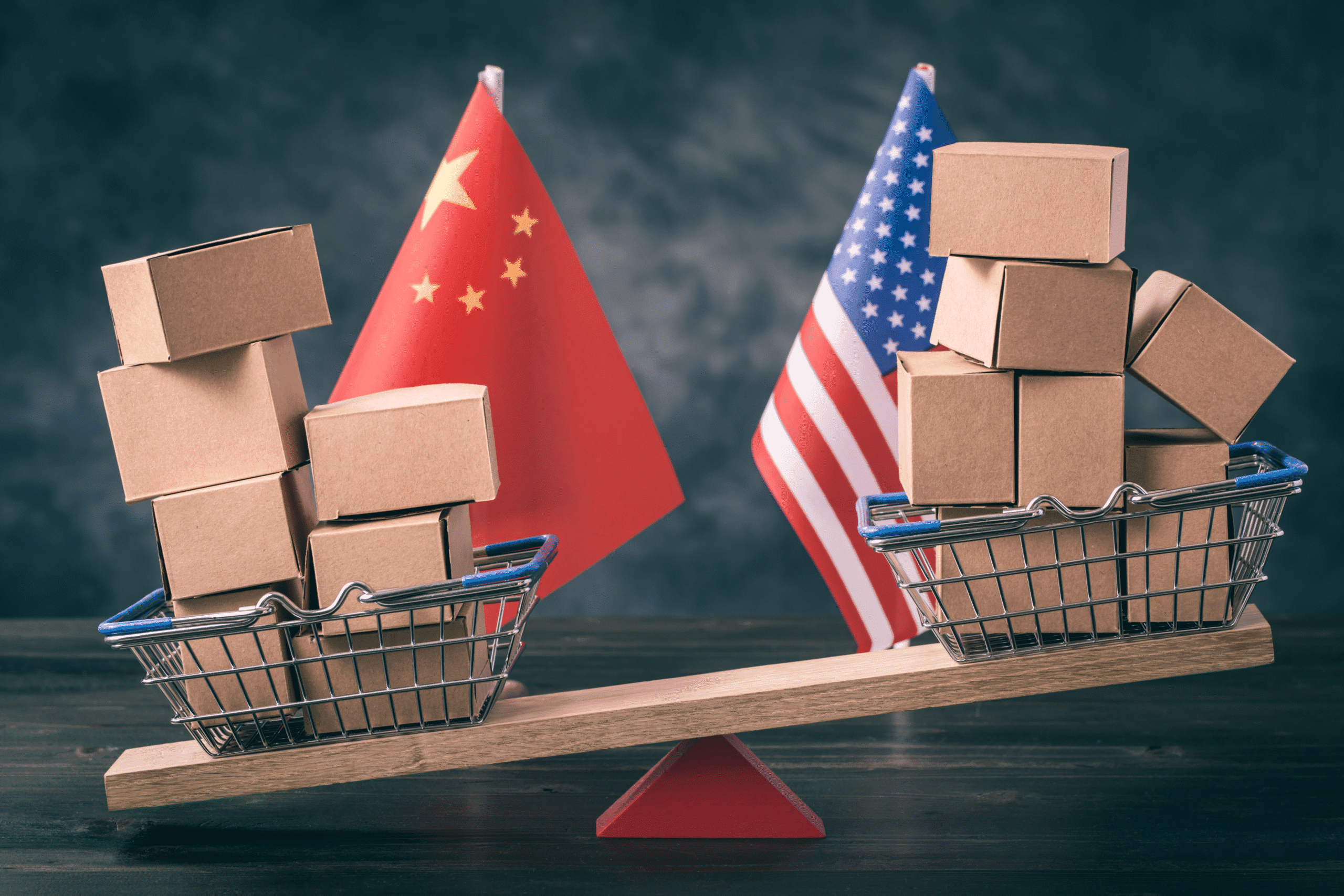 Cardboard boxes in shopping baskets sit either side of a wooden seesaw, placed in front of American and Chinese flags. This visually represents the ongoing trade war.