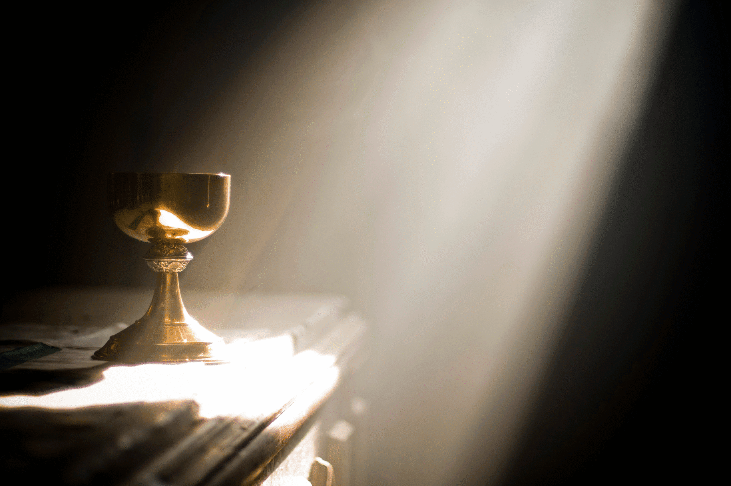 A golden chalice, presumably the Holy Grail, is sat on an altar and lit by a beam of natural light descending from above.