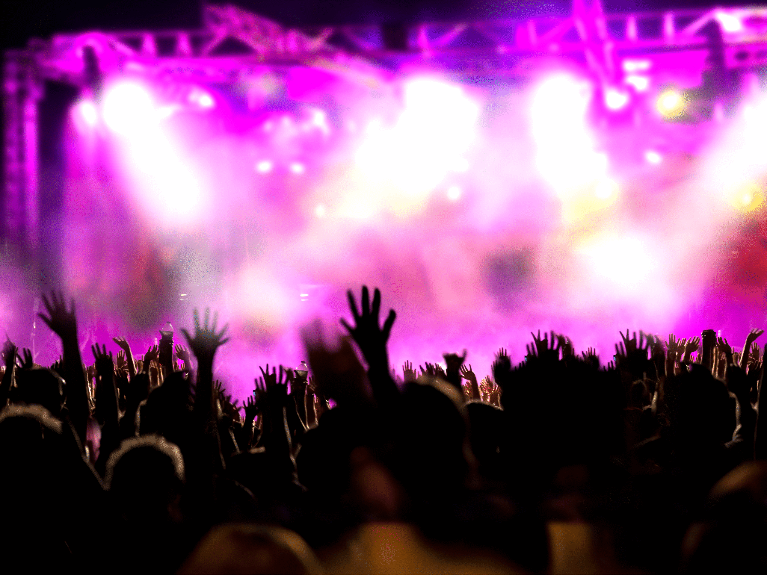 A live concert stage is lit in pink and purple light and covered in stage fog, while hundreds of fans raise their hands in the crowd.