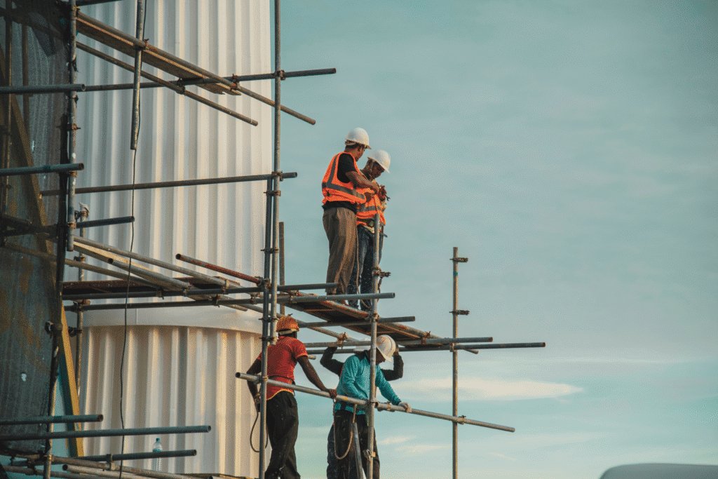 Two construction workers wearing hard hats and bright orange workers stood on scaffolding against a building