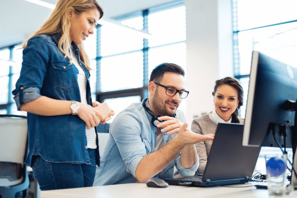 Three workers, two sitting and one standing, are looking at a laptop screen at an office desk.