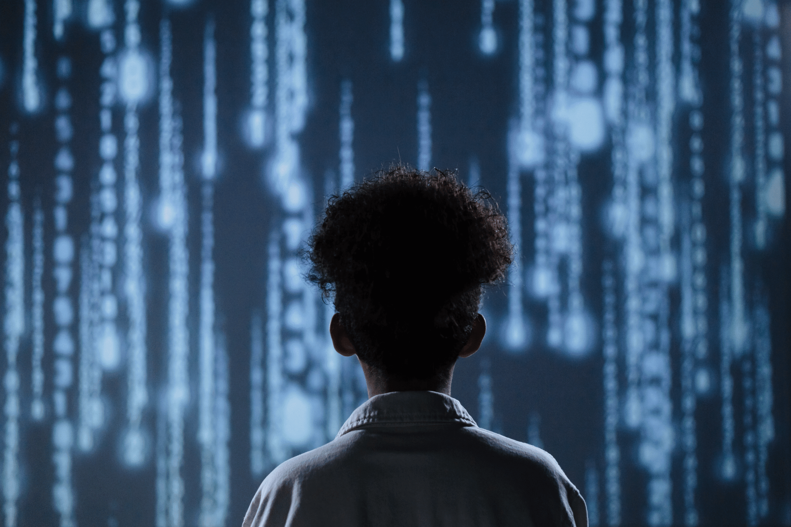 A man in a white shirt with dark curly hair has his back turned. He is facing a glowing wall of blue lights seemingly depicting computer code or AI software routines.