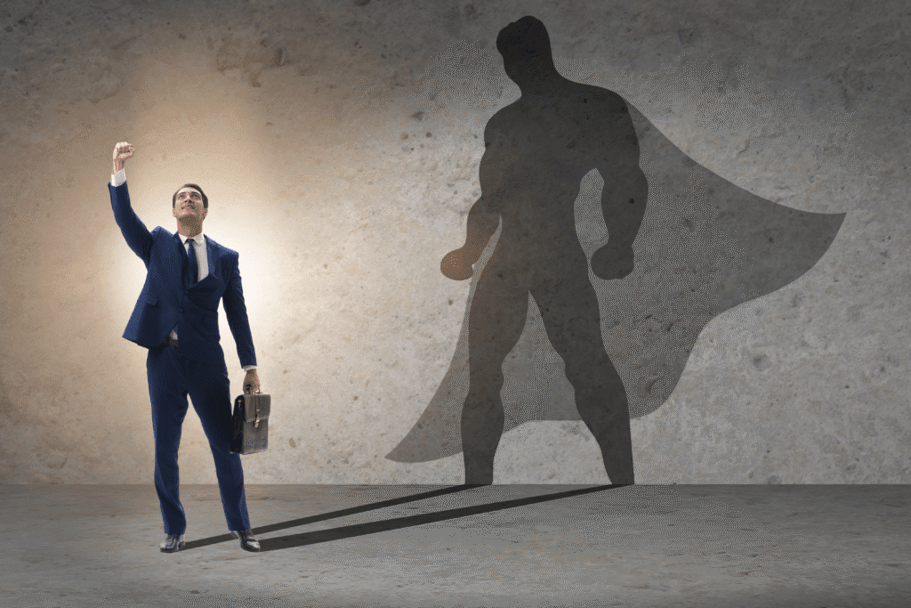 A businessman in a suit stands in front of a dark grey wall with his fist pointed up in the air. A Superman-like silhouette is projected behind him.
