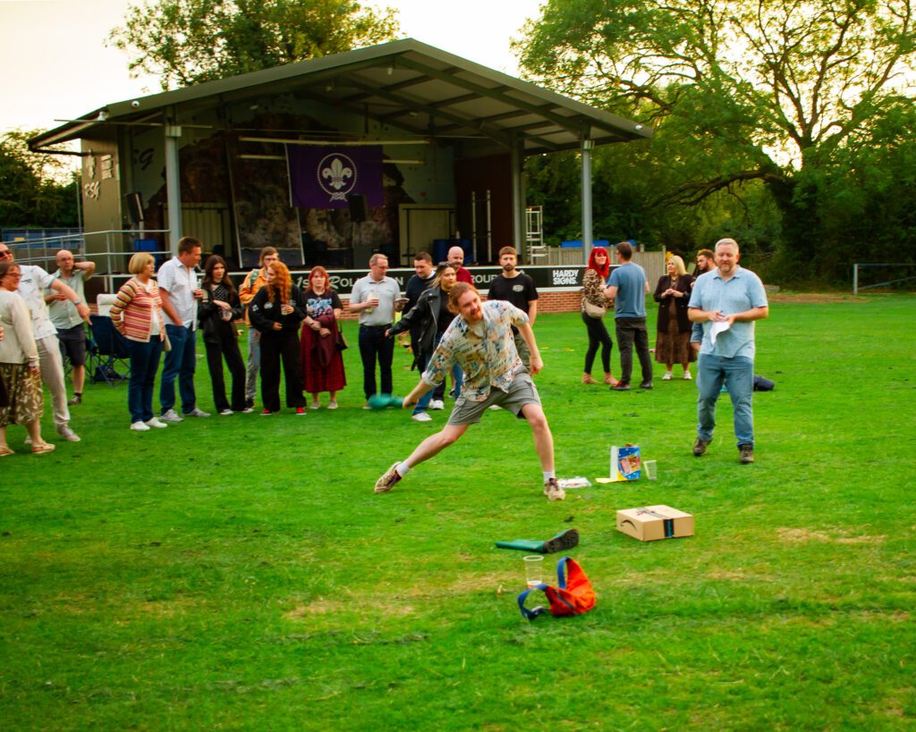 An action shot of a Solutions employee playing welly wanging, leaning down to throw the welly. Other Solutions employees are standing in the background watching.