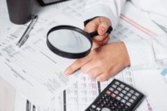 A close-up shot of an auditor's hands looking at paperwork on a desk, with a magnifying glass in one hand. 