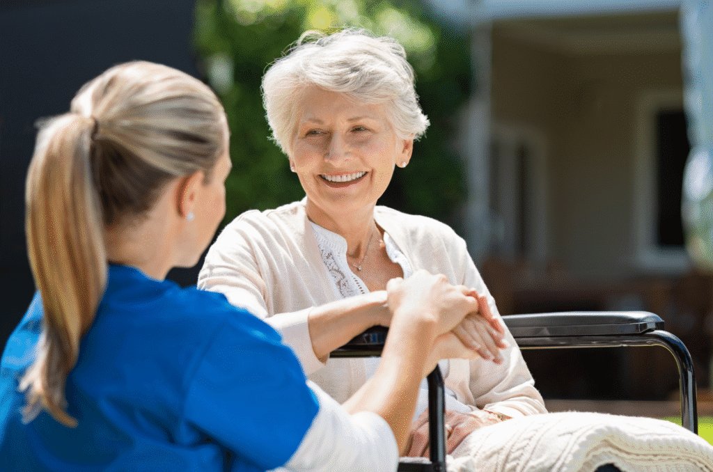 A close-up shot from behind of a care worker holding the hand of an elderly lady, who is sitting in a wheelchair and facing the camera.