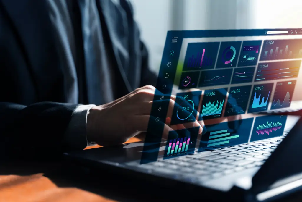 A close-up shot of an employee sitting at a desk using their laptop. There’s a futuristic business dashboard interface above it showing AI data.