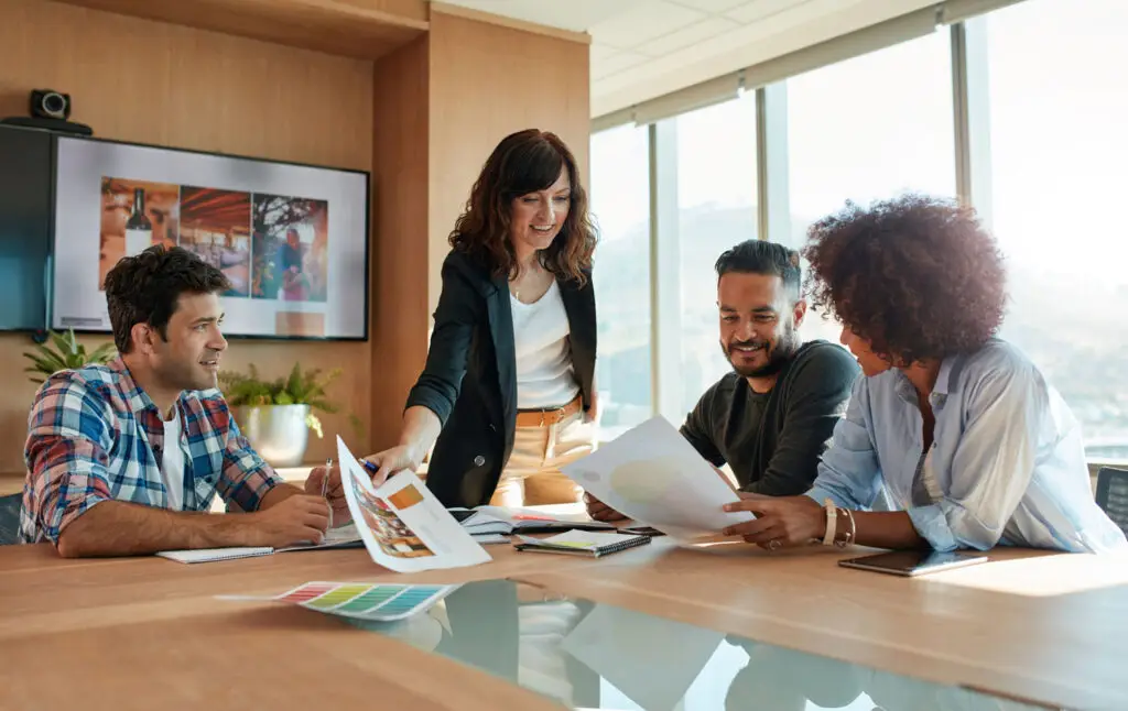 Four casually dressed people in a office building meeting around a table discussing documents