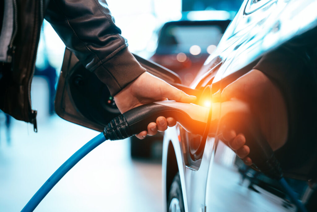 Close-up photo of a man plugging in a charger to his electric car, with a very saturated blue image and an orange light emitting from the charger port