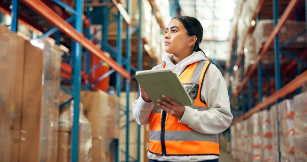 A woman is holding a tablet while standing in a warehouse, likely checking stock or conducting a quality assurance check.