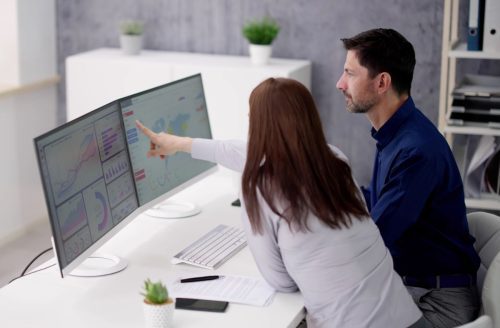 Two office workers sat at a desk, looking at data on the two monitors.